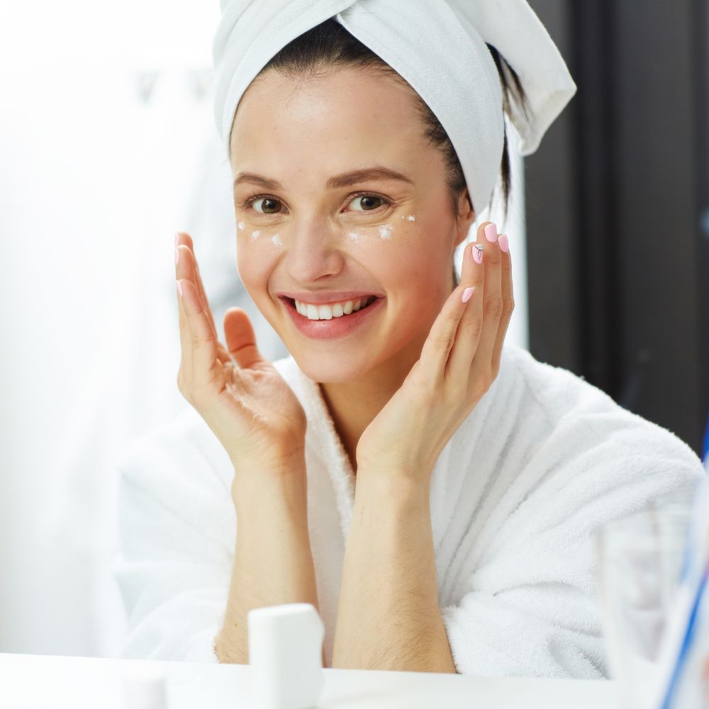 Happy woman in bathrobe applying moisturizer cream to glowing face